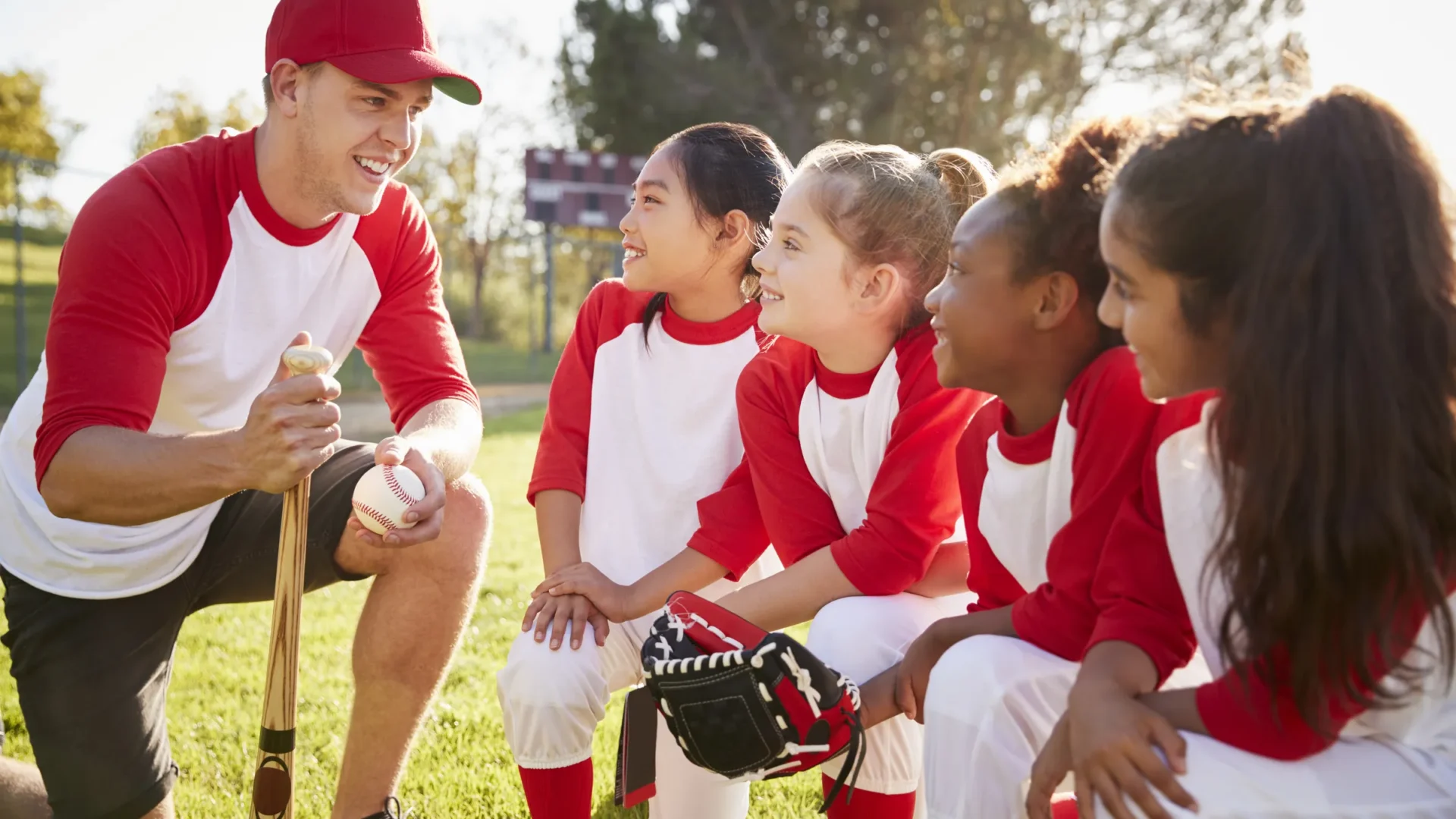 A smiling coach kneeling with his girls baseball team before a game